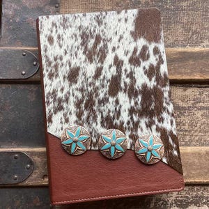 May include: A rectangular notebook featuring a cover with a brown and white cowhide pattern on the top half and solid brown leather-like material on the bottom half. Three circular metal conchos with turquoise star-shaped inlays are aligned.