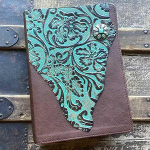 May include: A rectangular notebook with a brown cover featuring a large, triangular-shaped inlay of turquoise leather with a dark brown embossed floral pattern. A circular metal concho button with a turquoise center secures the decorative inlay.