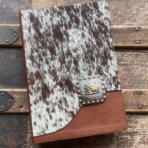 May include: A rectangular notebook cover featuring a brown and white cowhide pattern on the upper portion and solid brown leather on the lower section. A silver-toned metal concho with a gold-colored horse and cross emblem is centered.