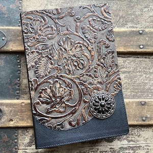 May include: A rectangular notebook featuring a dark brown cover with an embossed floral pattern and a smooth black leather-like bottom section. A circular black decorative button with a metallic rim is attached to the lower right corner.