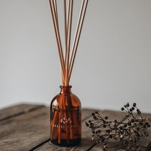 May include: A brown glass diffuser bottle with wooden sticks and the text "Embers + Roots" on the label. The bottle is sitting on a wooden surface with dried flowers.