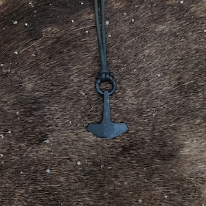 Puede incluir: Un collar con un colgante de metal negro en forma de martillo de Thor, llamado Mjolnir, en un cordón de cuero negro.