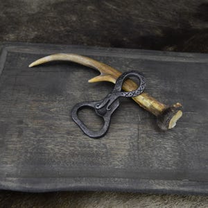May include: A hand-forged bottle opener with a decorative handle, resting on a section of deer antler. The opener is dark gray and lies on a dark wooden tray. The antler is light brown and beige.