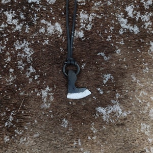 Könnte beinhalten: Ein kleiner schwarzer Axt-Anhänger mit einer silbernen Klinge hängt an einem schwarzen Lederband. Der Anhänger befindet sich auf einem braunen, pelzigen Hintergrund.