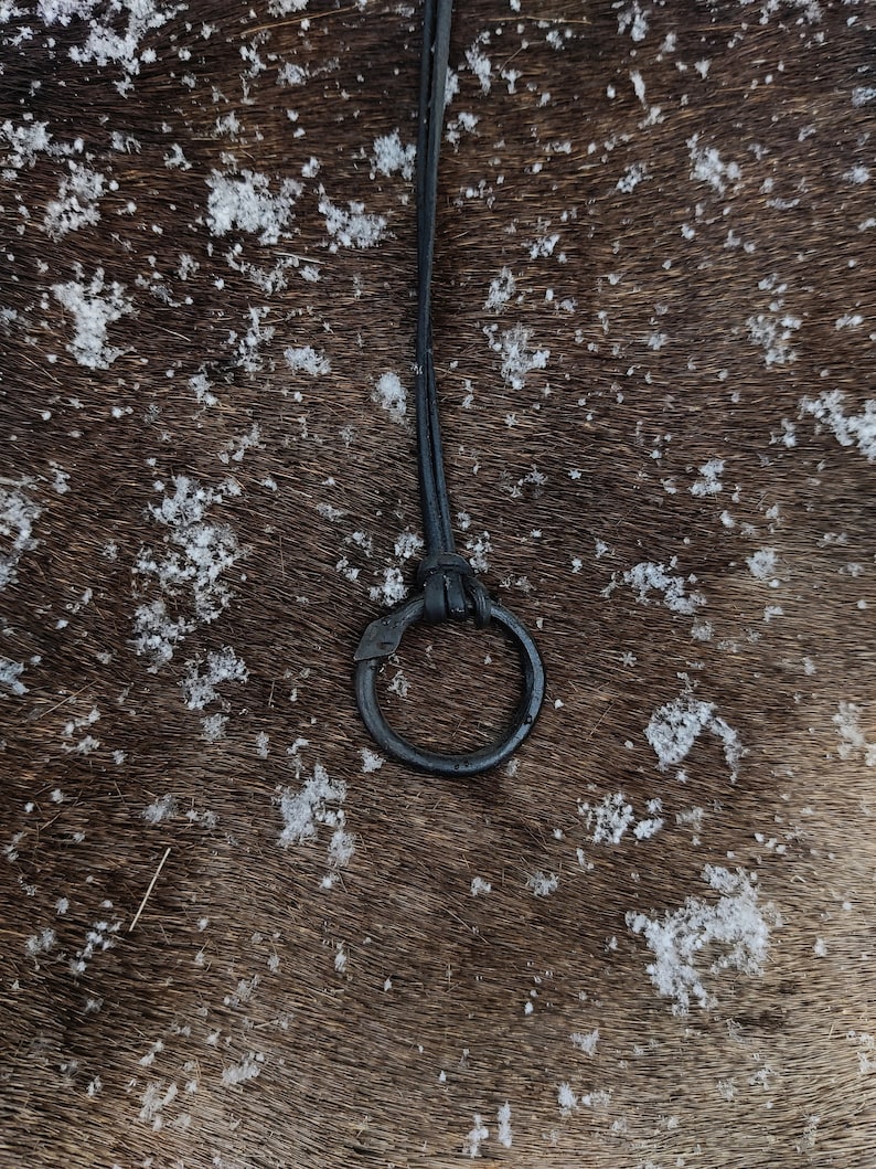 May include: A black leather strap with a metal ring attached to it. The strap is lying on a brown surface with white specks.
