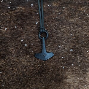 May include: A black leather cord necklace with a black metal pendant shaped like a hammer with a rune symbol on it. The necklace is on a brown fur background.
