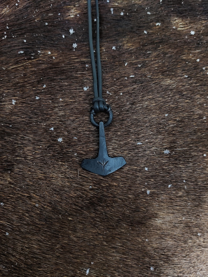 May include: A black leather cord necklace with a black metal pendant shaped like a hammer with a rune symbol on it. The necklace is on a brown fur background.