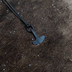 May include: A black leather cord necklace with a small, black metal hammer pendant. The pendant is a simple design with a rounded head and a flat, rectangular blade.