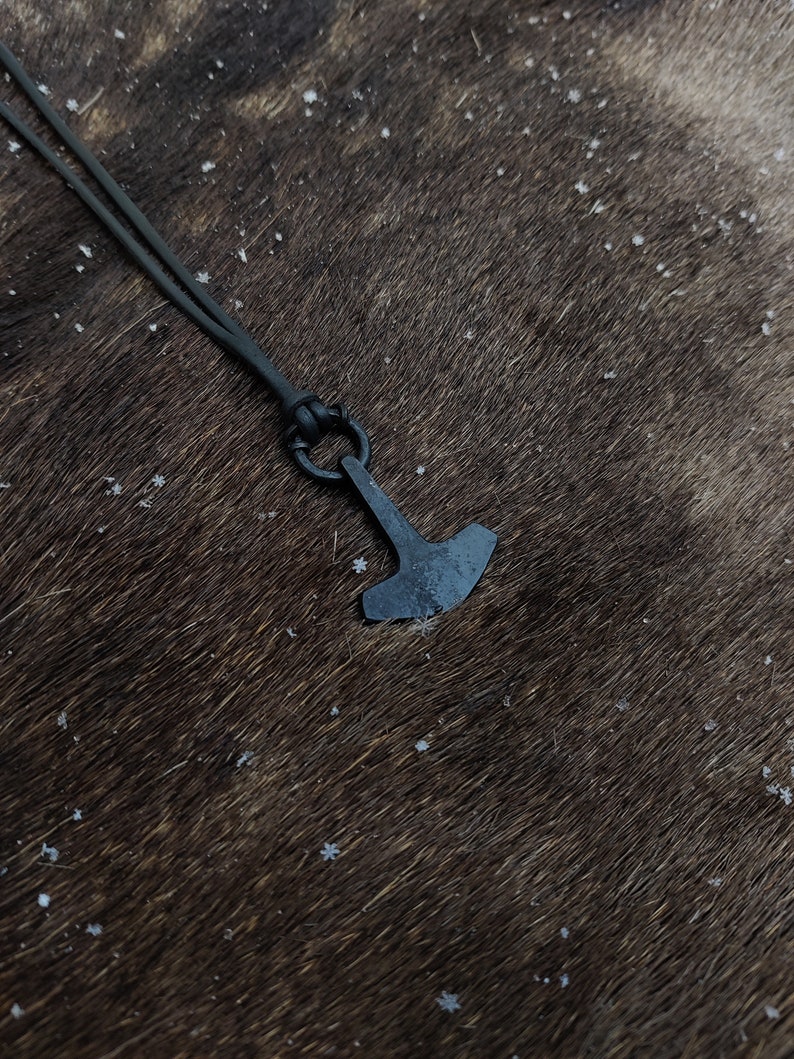 May include: A black leather cord necklace with a small, black metal hammer pendant. The pendant is a simple design with a rounded head and a flat, rectangular blade.