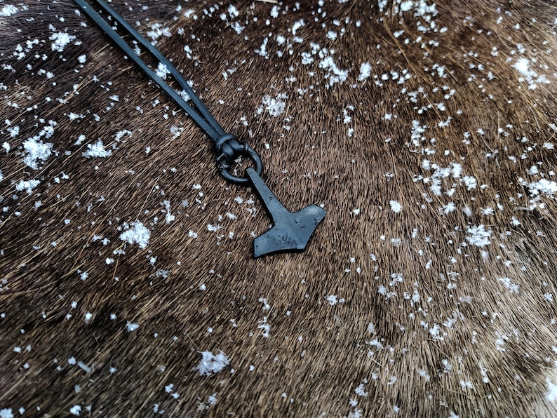 Pode incluir: Um colar de cord&atilde;o de couro preto com um pingente de martelo de metal preto. O colar est&aacute; sobre uma superf&iacute;cie peluda marrom com manchas brancas.