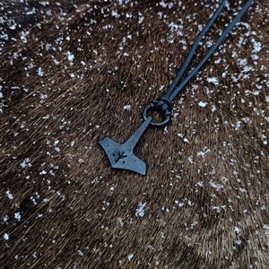 Pode incluir: Um colar de cord&atilde;o de couro preto com um pingente de metal preto em forma de martelo com um s&iacute;mbolo de runa gravado nele. O colar est&aacute; sobre uma superf&iacute;cie peluda marrom.