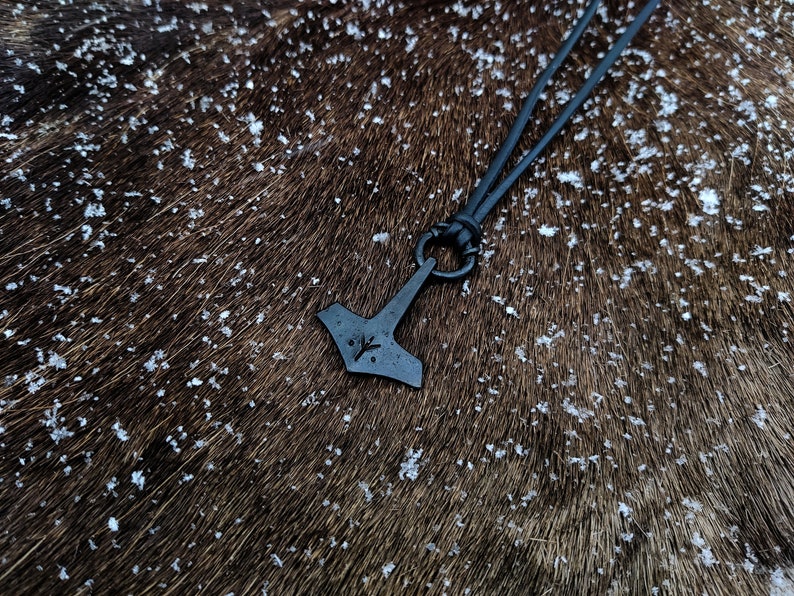 Pode incluir: Um colar de cord&atilde;o de couro preto com um pingente de metal preto em forma de martelo com um s&iacute;mbolo de runa gravado nele. O colar est&aacute; sobre uma superf&iacute;cie peluda marrom.
