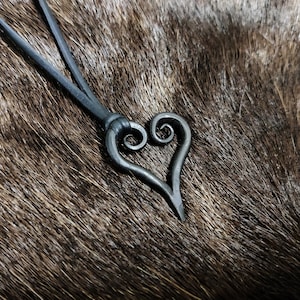 Op de afbeelding: Een ketting van zwart lederen koord met een handgesmeed hartvormig hanger. Het hart is van metaal en heeft een draaiend ontwerp.