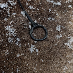 May include: A close-up of a black leather strap with a metal ring attached. The strap is lying on a brown surface with white specks.