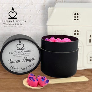 May include: A black round container with a white label that reads "La Casa Candles Wax Melts & Gifts Handmade in Devon Snow Angel 100% Soy Wax". The container is filled with pink heart-shaped wax melts with purple glitter.