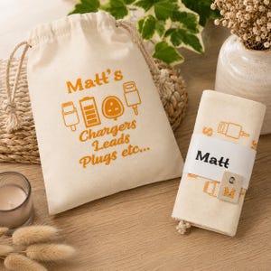 May include: A cream-coloured drawstring pouch with orange graphics of charging icons and the text Matt's Chargers Leads Plugs etc... next to a matching rolled fabric organiser featuring a white paper band labelled Matt and a small tag with the letter M.