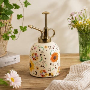 May include: A cream-coloured ceramic plant mister featuring a gold-toned metal pump mechanism. The body is decorated with a colourful pattern of sunflowers, small flowers, stars, crescent moons, and a bee. The pump has a ring handle and nozzle.