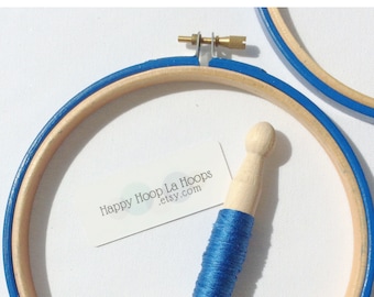 May include: A blue wooden embroidery hoop with a wooden embroidery needle and a spool of blue embroidery floss. The hoop is labeled "Happy Hoop La Hoops etsy.com".