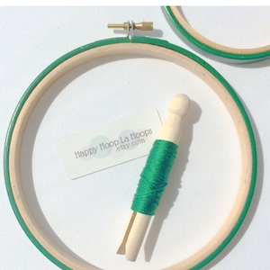 May include: A green embroidery floss wound around a wooden embroidery hoop with a green outer ring. The hoop is on a white surface with a business card that reads "Happy Hoop La Hoops etsy.com".