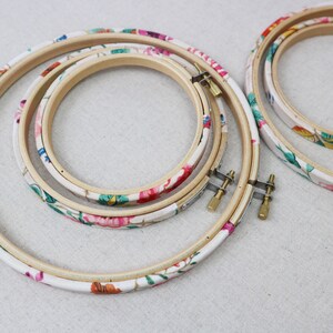 May include: Three embroidery hoops with floral patterned fabric covering the wooden hoops. The hoops are nested together, with the largest hoop on the bottom and the smallest hoop on top.