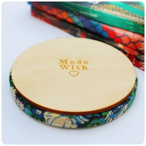 May include: A round wooden coaster with a floral fabric edge. The top of the coaster is made of light wood and has the words "Made With" and a heart engraved on it.