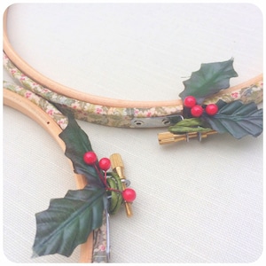 May include: Two embroidery hoops with green holly leaves and red berries attached to them. The hoops are wrapped in floral patterned fabric.