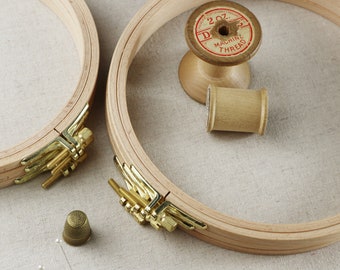 May include: Two wooden embroidery hoops with gold metal clasps. A spool of thread with the label "2 oz. D.M. Machine Thread" is in the center of the image. A thimble and sewing pins are on the table.