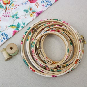 May include: A set of six embroidery hoops with floral patterns in shades of pink, green, and yellow. The hoops are nested inside each other, with the largest hoop at the bottom and the smallest hoop at the top.