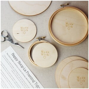 May include: Three wooden embroidery hoops with the text "Made With" and a heart symbol engraved on each. The hoops are on a beige fabric surface with a sheet of paper with instructions for a hoop-la framing tutorial.