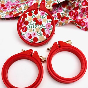 May include: Three red embroidery hoops, two with a plain red finish and one with a white fabric background with a red and pink floral pattern.