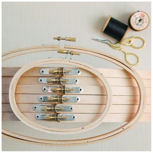 May include: Several wooden embroidery hoops with brass fixings are displayed. A spool of black thread, a vintage thread spool, and gold scissors are also present, suggesting a crafting theme.