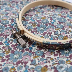 Könnte beinhalten: Ein hölzerner Stickrahmen mit einem Metallspannmechanismus liegt auf floralem Stoff. Der Stoff zeigt ein Muster aus kleinen blauen, lila und gelben Blumen auf cremefarbenem Hintergrund. Der Rahmen ist aus hellem Holz gefertigt.