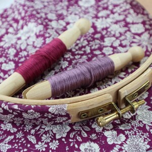 May include: Two wooden embroidery hoops with purple and pink embroidery floss wound around them. The hoops are on a purple and white floral fabric.