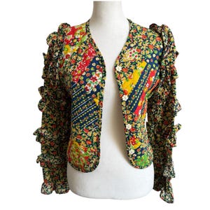 May include: A floral print jacket with a V-neck and ruffled sleeves. The jacket features a patchwork design with red, yellow, and blue floral patterns. It has a button closure and is displayed on a white mannequin.