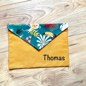 Può includere: Una custodia a forma di busta giallo senape con un risvolto turchese con un motivo tropicale di scimmie, tucani e fogliame. Il nome "Thomas" è stampato in nero sul davanti. La custodia è probabilmente per riporre piccoli oggetti.