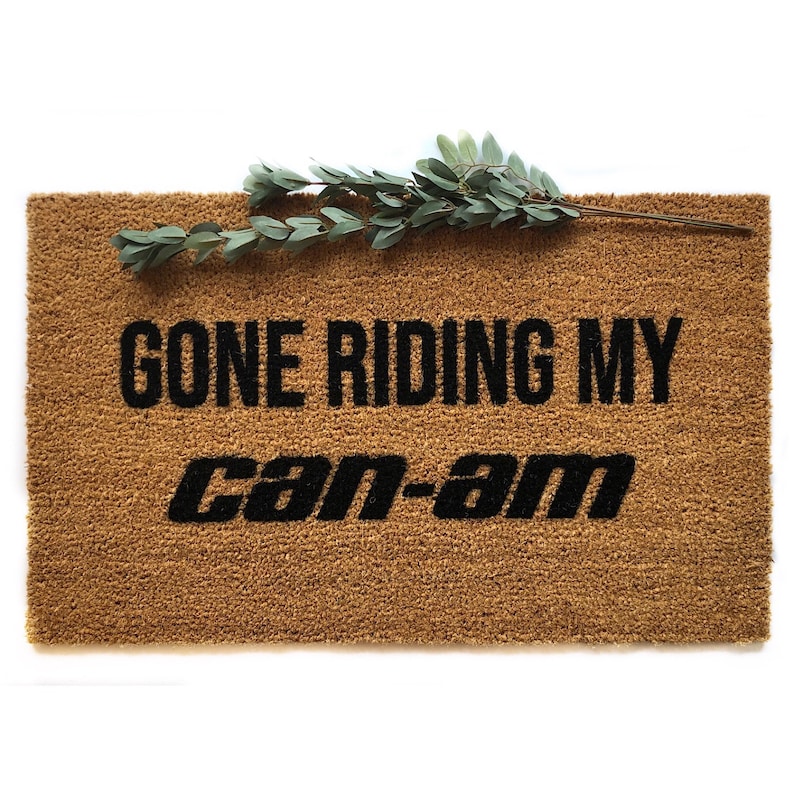May include: A brown coir doormat with the text "Gone Riding My Can-Am" in black lettering.