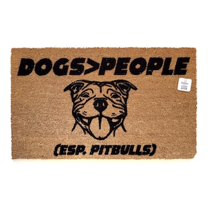 Peut inclure: Un paillasson en fibre de coco brun avec le texte "DOGS>PEOPLE (ESP. PITBULLS)" en noir. Une silhouette noire de la tête d'un pit-bull est centrée au-dessus du texte.