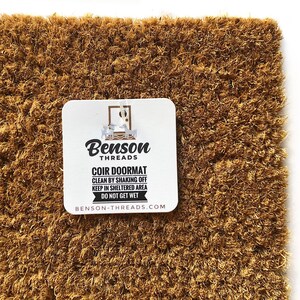 May include: A rectangular coir doormat with a natural brown color. A white tag with the text "Benson Threads COIR DOORMAT" is attached. The tag also includes cleaning instructions and the website address.