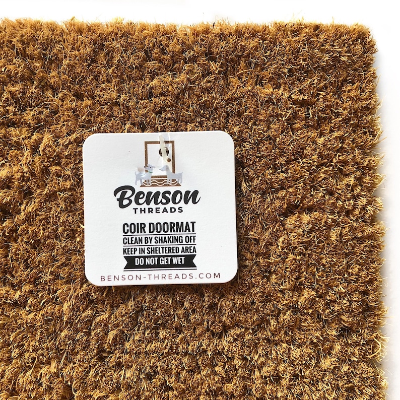 May include: A rectangular coir doormat with a natural brown color. A white tag with the text "Benson Threads COIR DOORMAT" is attached. The tag also includes cleaning instructions and the website address.