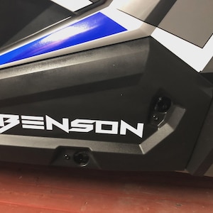 Könnte beinhalten: Ein Abschnitt eines schwarz-grauen Fahrzeugs mit einem weißen "BENSON"-Aufkleber. Ein blauer Streifen akzentuiert das Design. Die Seitenwand des Fahrzeugs hat ein geometrisches Muster.