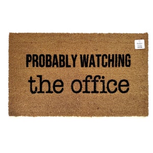 以下が含まれることがあります： 黒文字で「PROBABLY WATCHING the office」と書かれた茶色のココナッツ繊維のドアマットです。