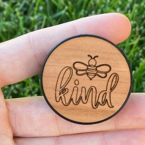 May include: A round wooden accessory with a black border. The center features a laser-etched bee and the word "kind" in a cursive font. The wood has a light brown color.