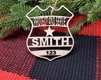 May include: Wooden police badge ornament with the words "PROTECT AND SERVE", a star, the name "SMITH", and the number "123". The ornament is hanging from a twine string. The background is a red and black plaid pattern.