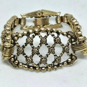 May include: A gold-toned bracelet with a delicate, intricate design. The bracelet features a series of small, round beads and a central section with a lattice pattern. The bracelet has a clasp closure.