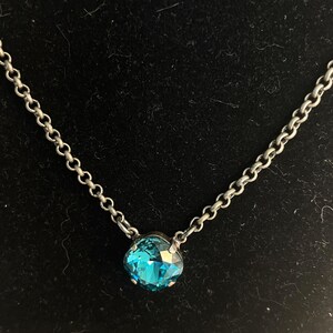 May include: A silver-toned necklace with a single, round, faceted, teal-colored gemstone pendant. The chain is made of small, interconnected links. The pendant is set in a silver-toned metal. The background is black.