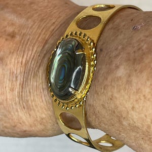 May include: Gold-tone cuff bracelet featuring an oval, iridescent stone with swirling patterns of green, blue, and gray. The bracelet has an open design with decorative cut-out ovals and a textured border around the stone.