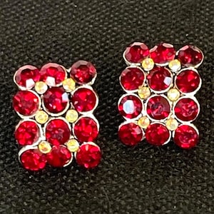 May include: A pair of square-shaped stud earrings. Each earring features a grid of vibrant red, round gemstones, interspersed with smaller, iridescent yellow stones. The earrings are set against a dark background.