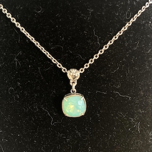 May include: A silver-toned necklace with a square, light green pendant. The pendant is set in a silver frame and hangs from a delicate chain. The background is a dark, textured surface.