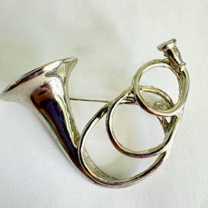 May include: A silver-toned brooch in the shape of a French horn. The brooch features a flared bell, a curved body, and a series of circular accents. The pin is designed to be worn on clothing or accessories.