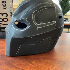 May include: A black, angular helmet prototype with white markings and an eye opening. The helmet is made of a matte material and sits on a wooden surface. The design features multiple panels and curved lines.